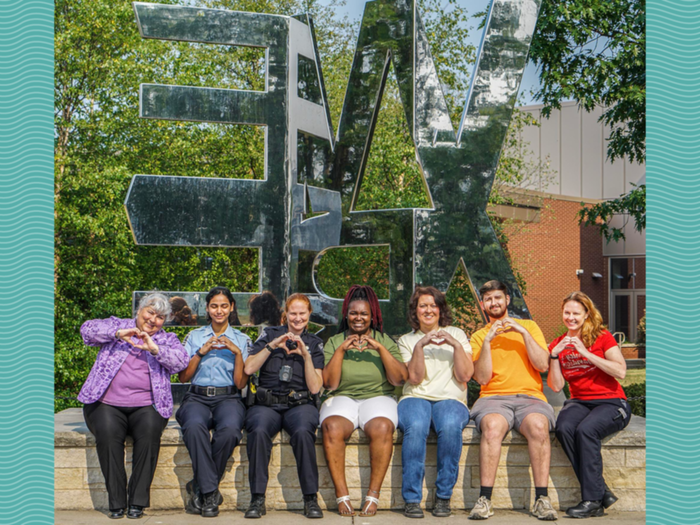 People in rainbow colored clothing sit in front of "We Are" statue with heart hands.