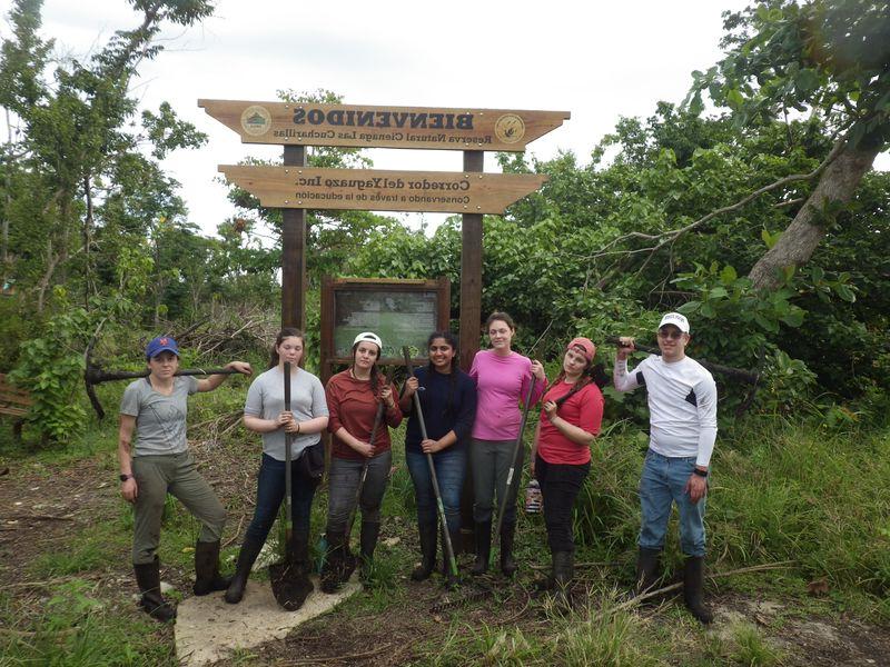 Seven students pose under a sign that says "Bienvenidos: Reserva Natural Cienaga Las Cucharillas"
