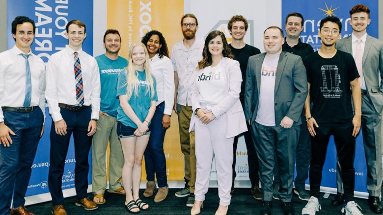 12 students pose standing for photo in front of Invent Penn State signage