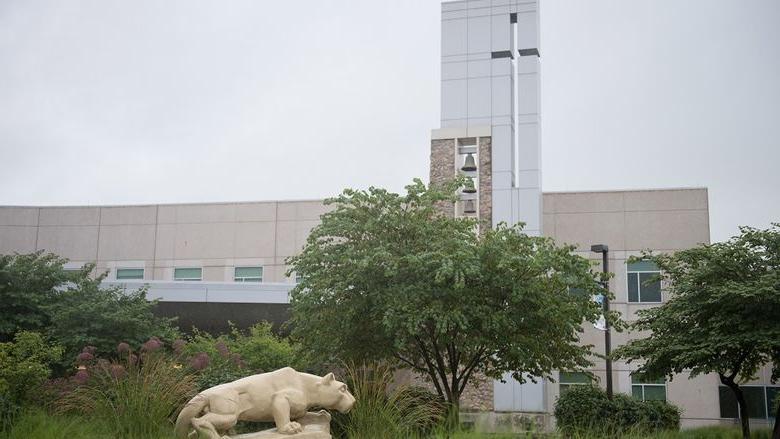 A statue of the Penn State Nittany Lion at the Penn State Health St. Joseph campus appears in profile in front of a row of trees and weeds. Behind the scene rises a glass tower and bells and windows.