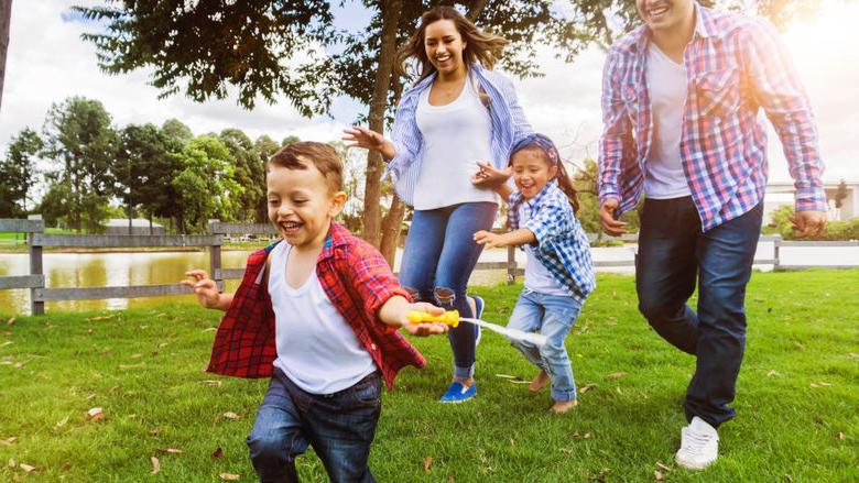 A man, a woman, and two children smile as they run through a park. trees and a fence are in the background.