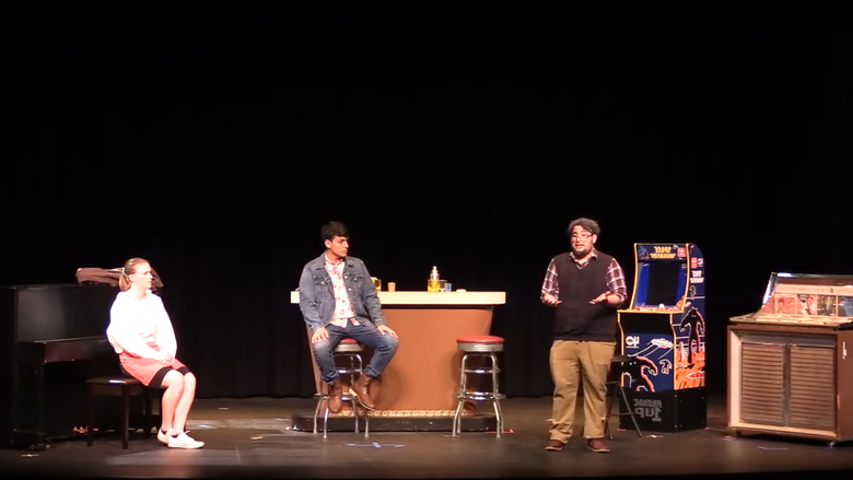 two men are sitting at a bar, one man talking animatedly, and a woman is sitting backward at a piano bench listening