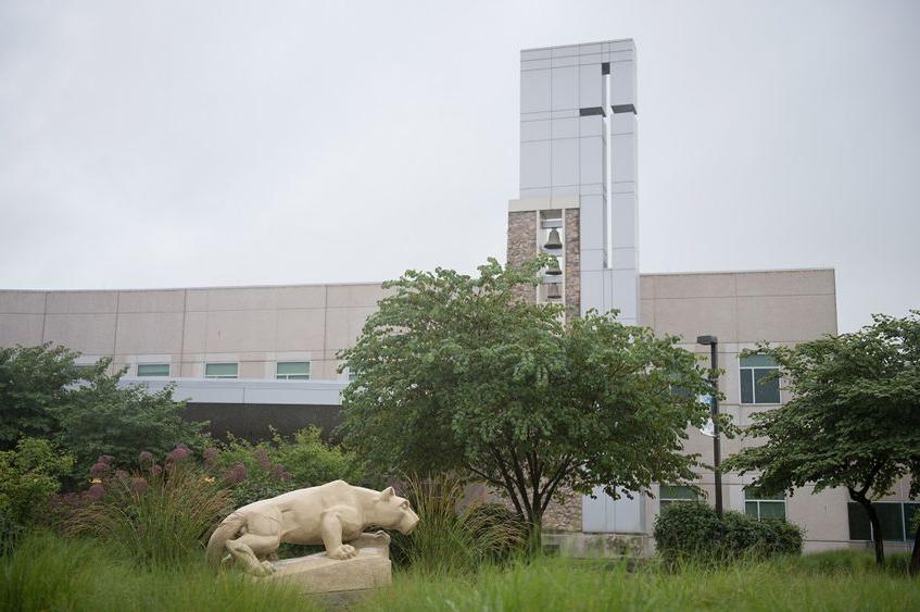 A statue of the Penn State Nittany Lion at the Penn State Health St. Joseph campus appears in profile in front of a row of trees and weeds. Behind the scene rises a glass tower and bells and windows.
