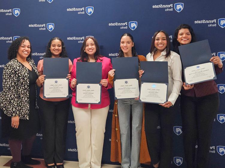Five students hold certificates open with a staff member, all standing in front of a branded Penn State banner