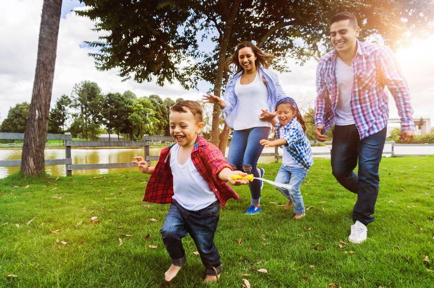 A man, a woman, and two children smile as they run through a park. trees and a fence are in the background.
